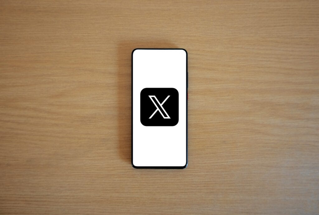 Minimalist image of smartphone with X logo on wooden surface, showcasing digital branding.